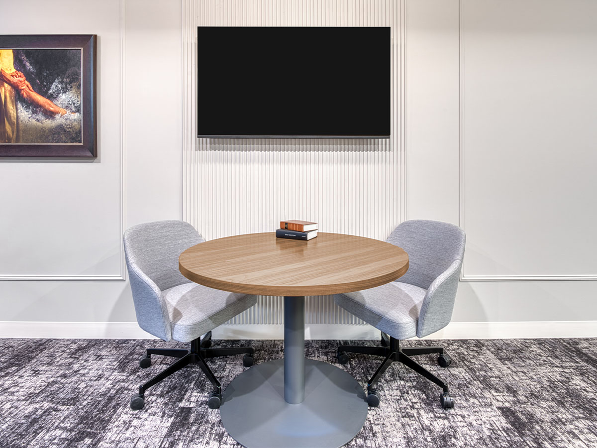 Round wooden table with two gray office chairs on a patterned carpet beneath a blank wall-mounted TV.