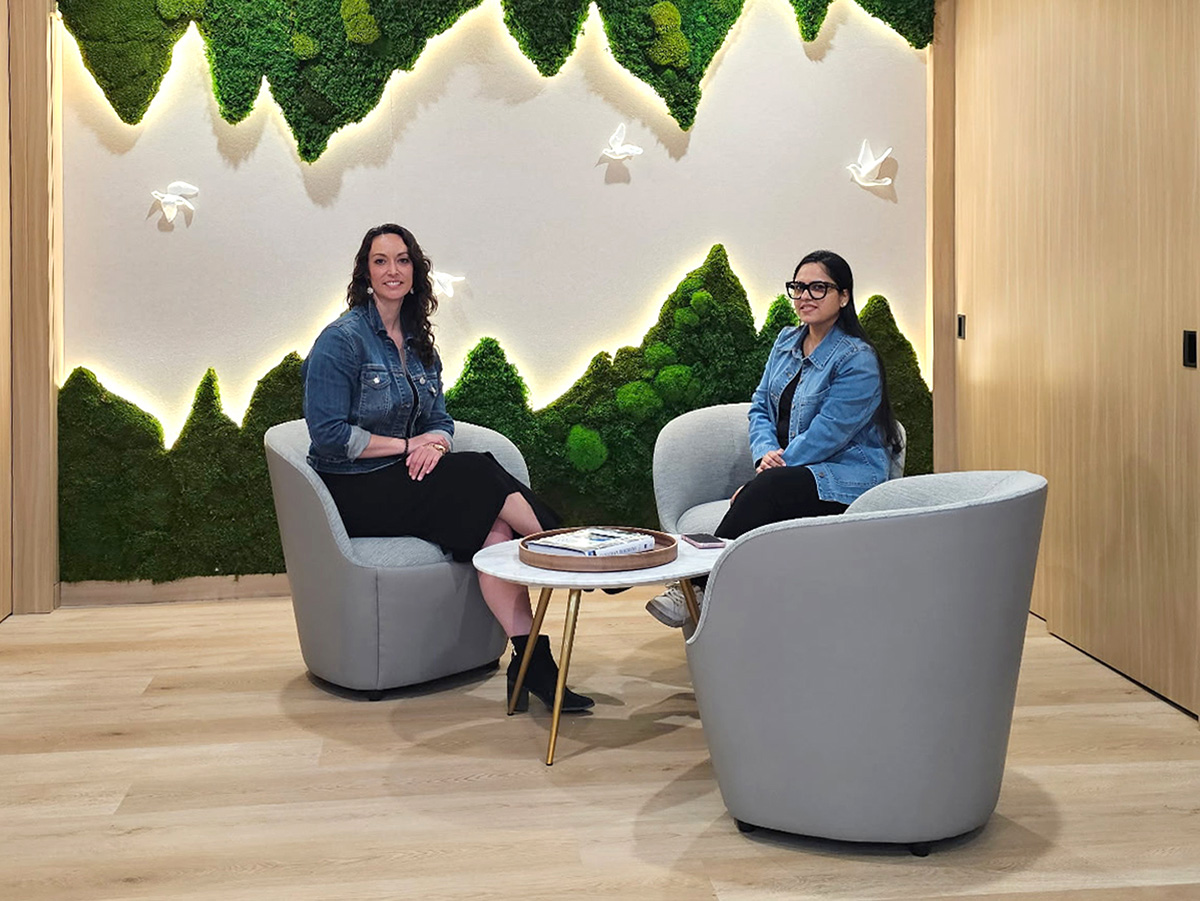 Two women sitting on gray chairs around a small table with plants and bird wall decor behind them.