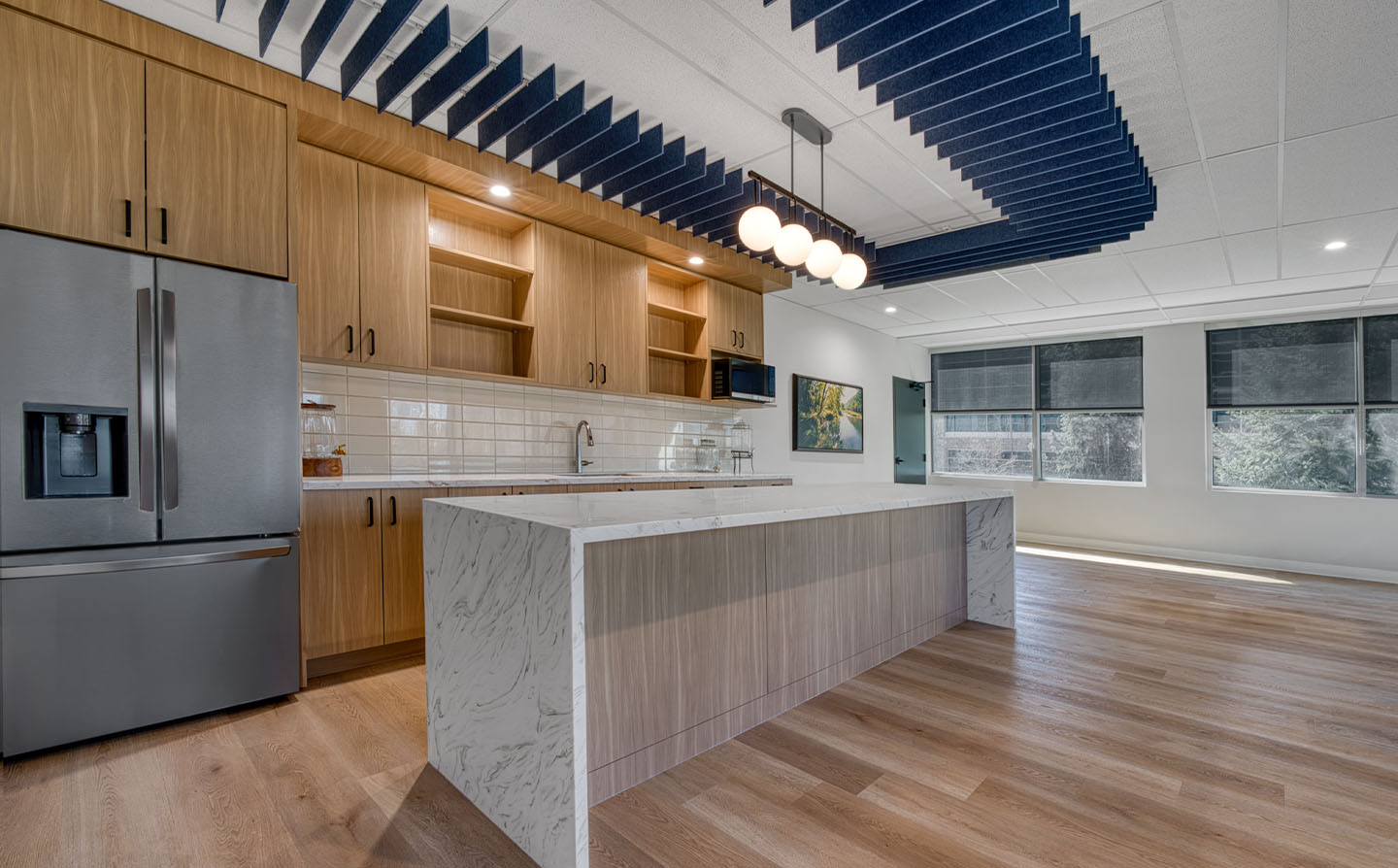 Office kitchen with wood cabinetry, marble island, globe pendant lights, and navy acoustic ceiling baffles.