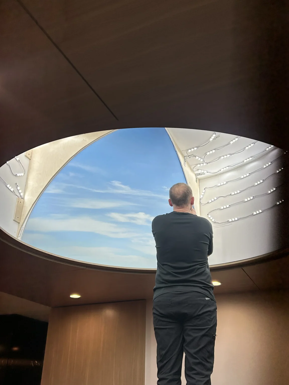 Man in black clothes standing indoors looking up at a large, triangular skylight with blue sky and clouds outside, with white string lights decorating the skylight frame.