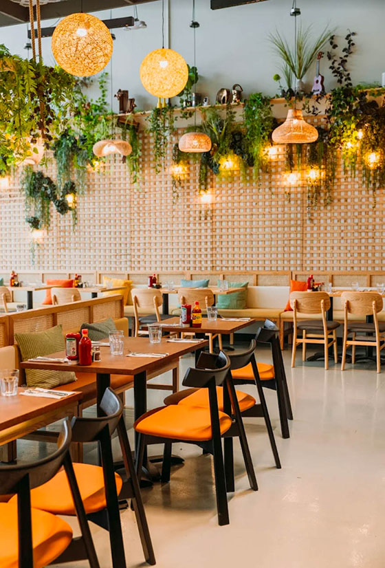 Bright, leafy bistro dining room with warm wood tables, woven pendants, and vibrant accent seating for a relaxed, inviting feel.