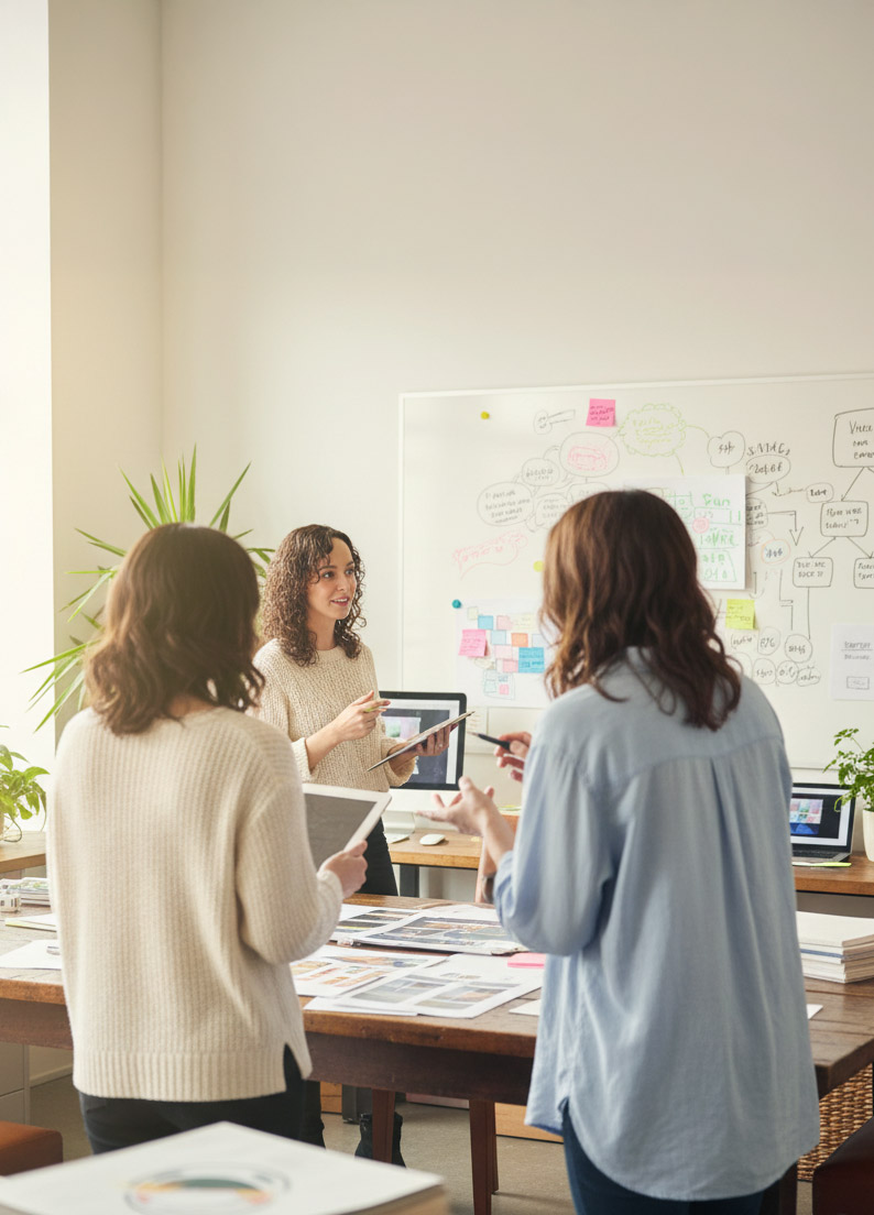 Three women discussing project materials at a shared office table.