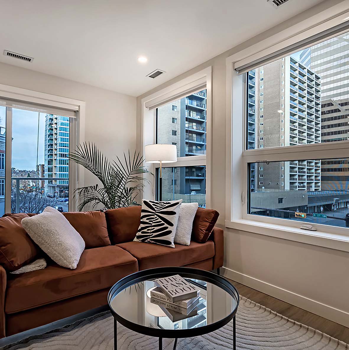 Sunlit corner living room with warm leather sofa, layered textures, and city views through oversized windows.