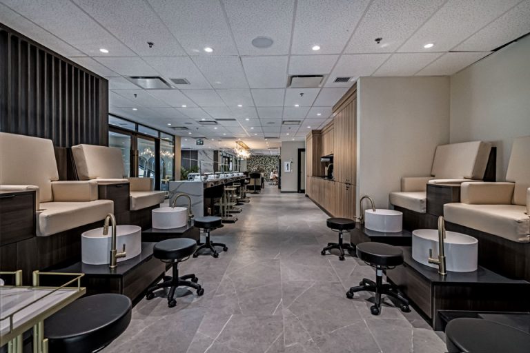Modern spa pedicure room with paired cushioned chairs, white basins, black stools, long marble stations, and recessed lighting.