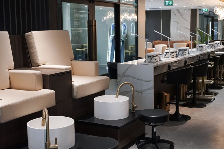 Modern spa pedicure area with marble stations, cushioned chairs, brass faucets, swivel stools, and sleek glass partitions.