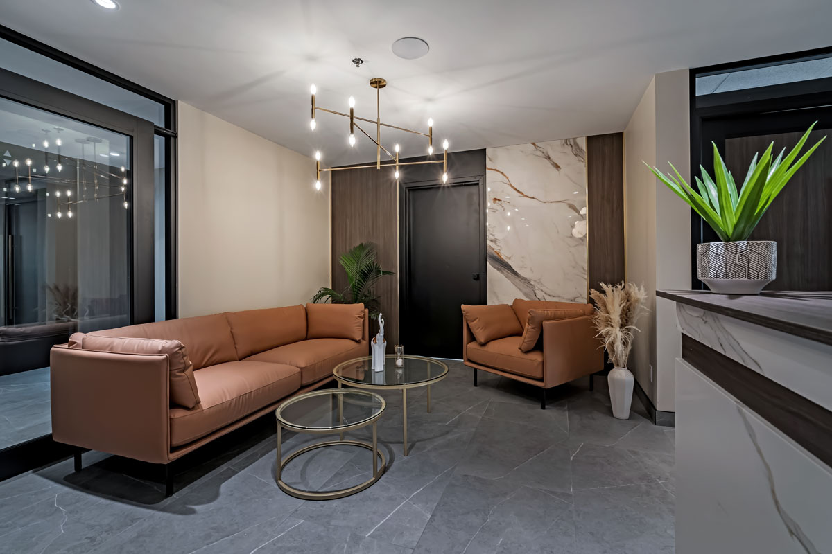 Modern spa reception with tan seating, round glass tables, marble accent wall, brass chandelier, and sleek front desk.