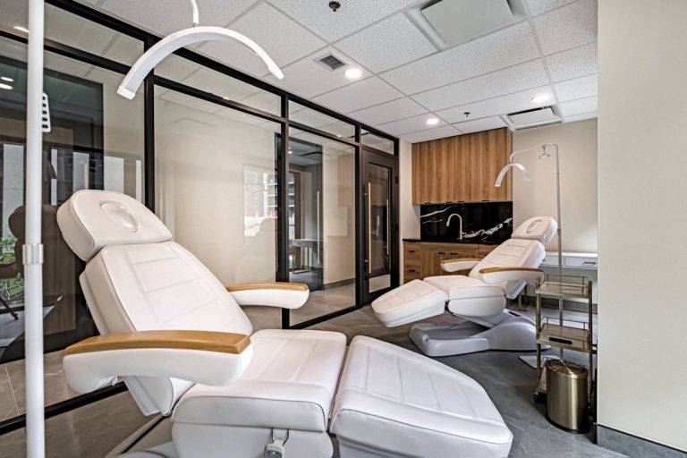 Modern spa treatment room with two adjustable reclining chairs, overhead lights, glass walls, wood cabinetry, and clean minimalist design.