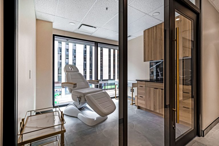 Modern spa treatment room with adjustable lounge chair, city-view windows, glass partition, wood cabinetry, and clean minimalist finishes.