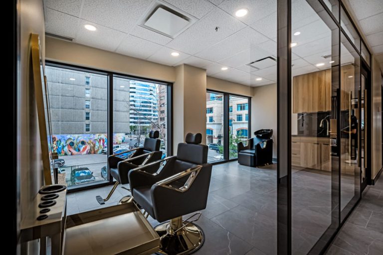Modern hair salon with styling chairs, floor-to-ceiling windows, city view, glass-partitioned rooms, and sleek lighting.