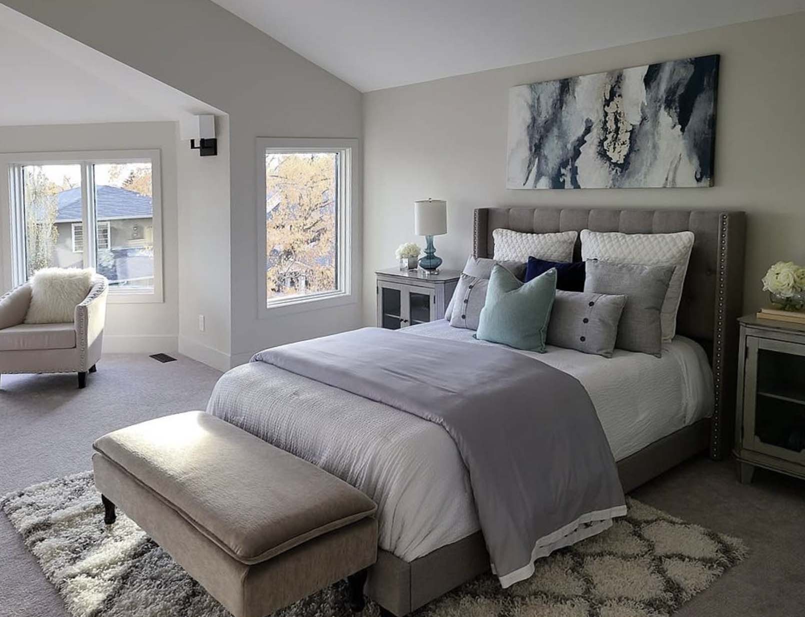 Modern bedroom with upholstered bed, neutral bedding, bench at foot, abstract wall art, side tables, and bright corner window.
