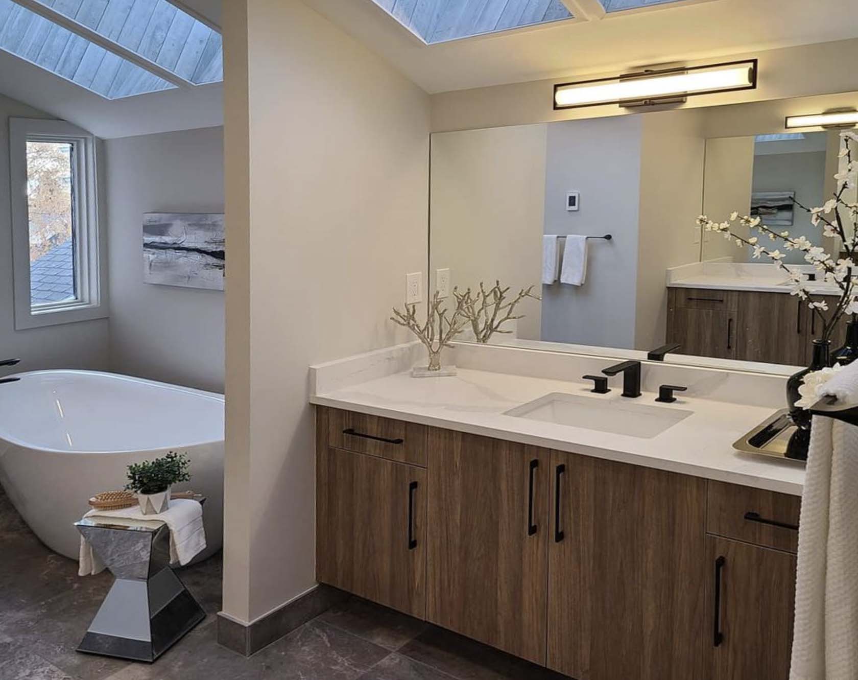 Modern bathroom with wood vanity, white countertop, black fixtures, skylit soaking tub, and neutral tile flooring.