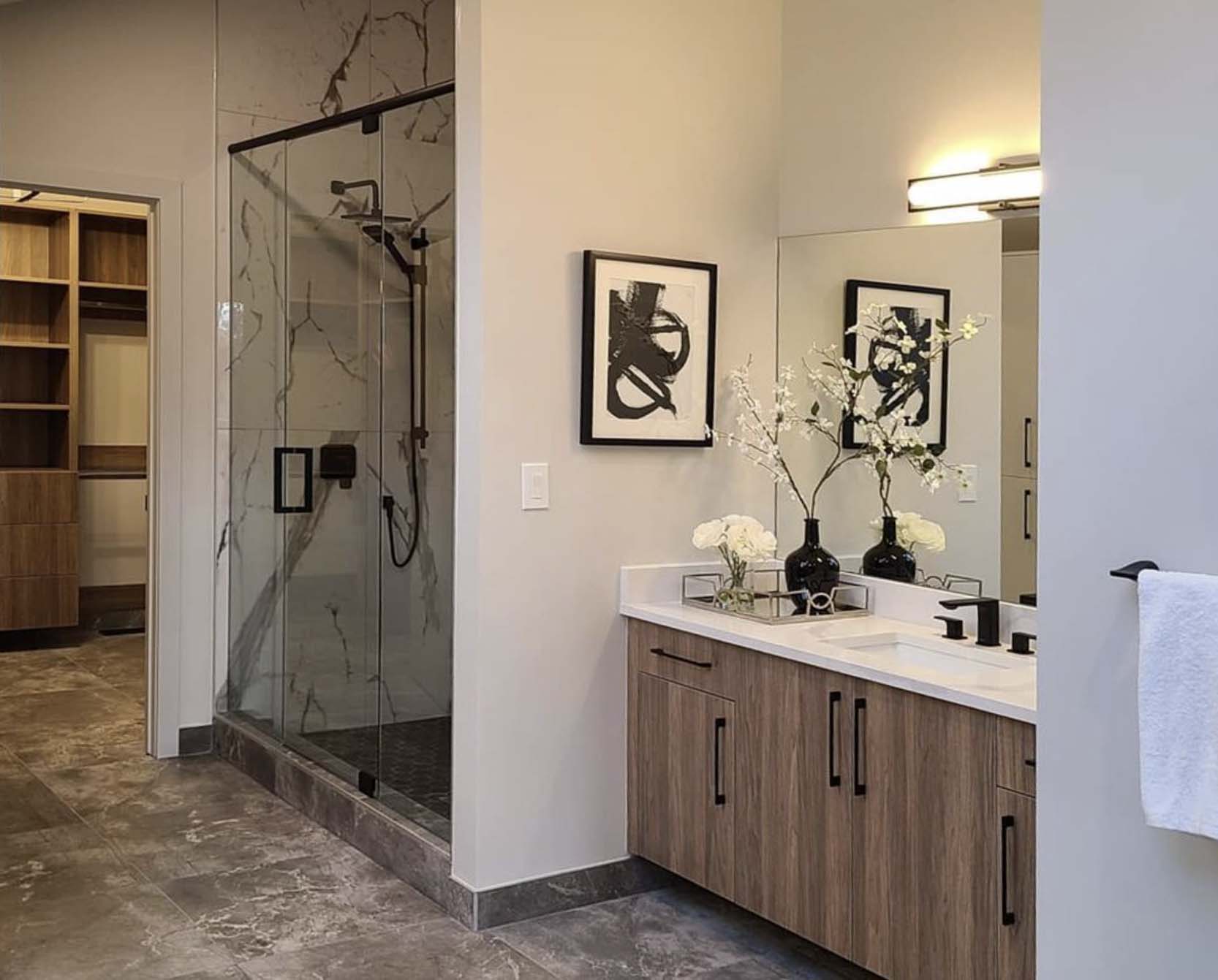Modern bathroom with wood vanity, white countertop, black fixtures, glass shower with marble tile, and framed abstract art.