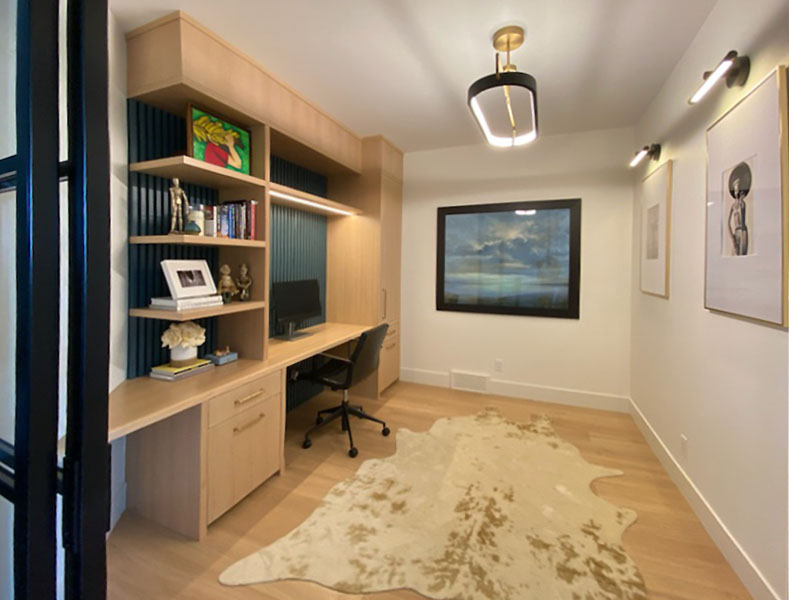 Modern home office with built-in wood desk and shelving, black slat accent wall, cowhide rug, framed art, and ceiling light.