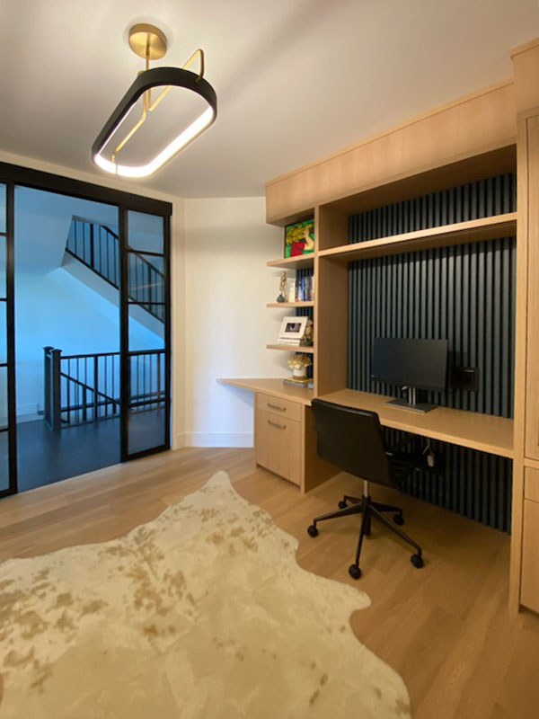 Modern home office with built-in wood desk, black slat wall, glass sliding doors, and contemporary ceiling light.