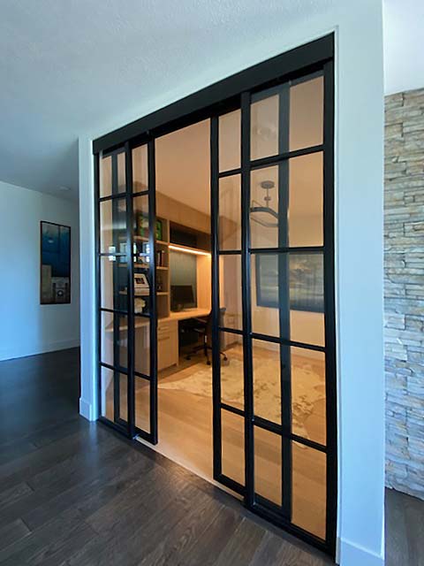 Modern home office behind black-framed glass sliding doors, with built-in desk, warm lighting, and dark wood floors.