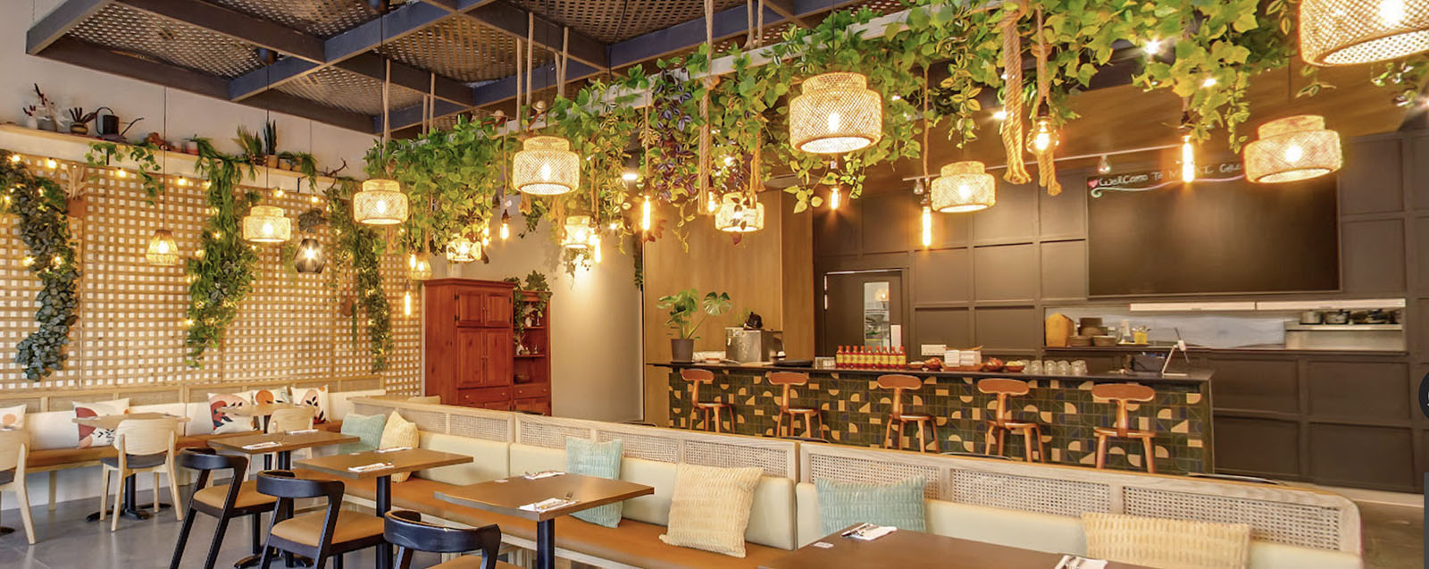 Warm, plant-filled restaurant with woven pendant lights, cozy banquettes, and a welcoming bar for relaxed dining.
