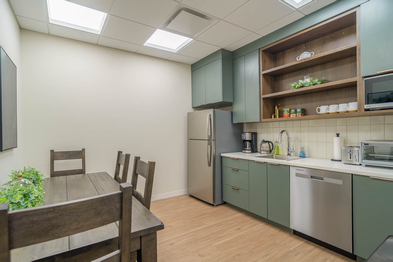 Staff kitchenette with sage cabinetry, open wood shelving, stainless appliances, and a compact dining nook.