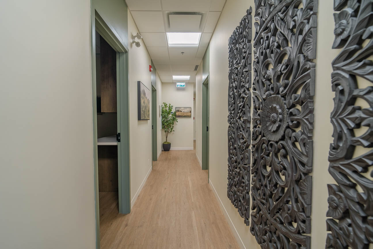 Warm, light-filled corridor with natural wood floors, calming sage doors, and carved wall art for a welcoming clinic experience.