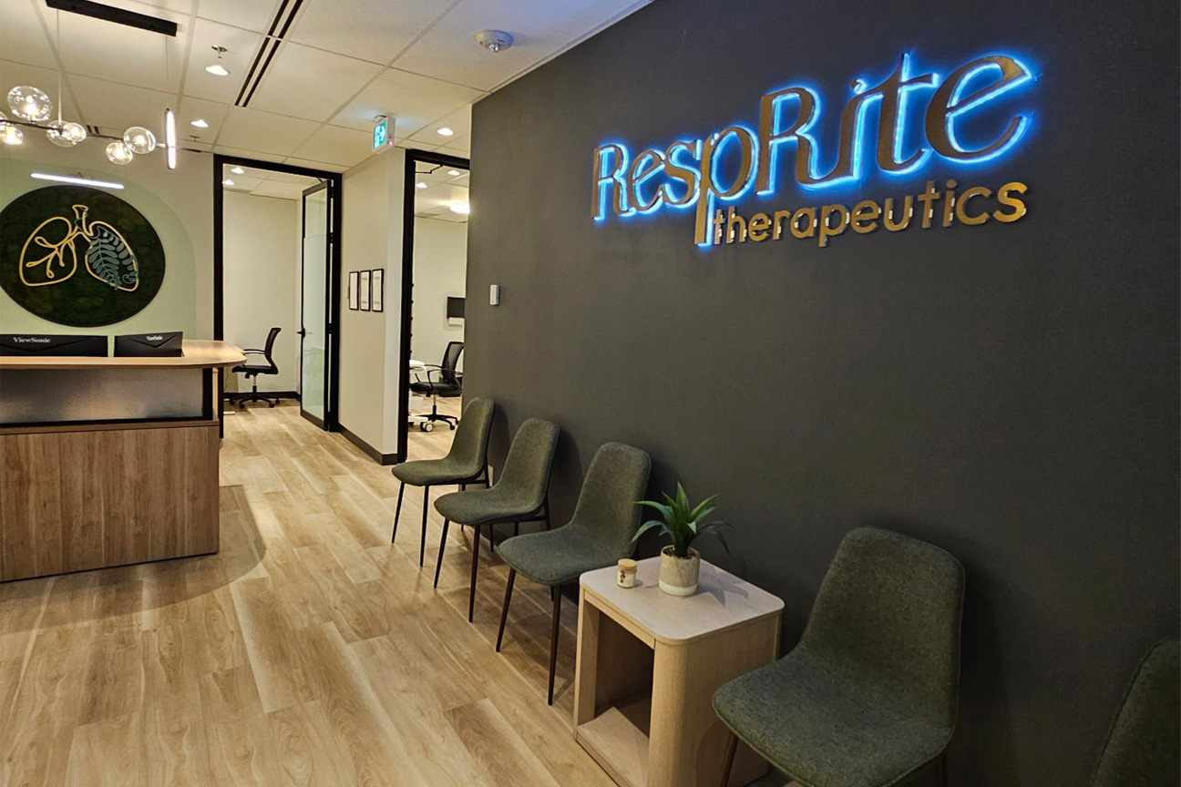 RespRite Therapeutics reception area with illuminated logo sign, wood desk, moss artwork, globe pendants, and grey waiting chairs.