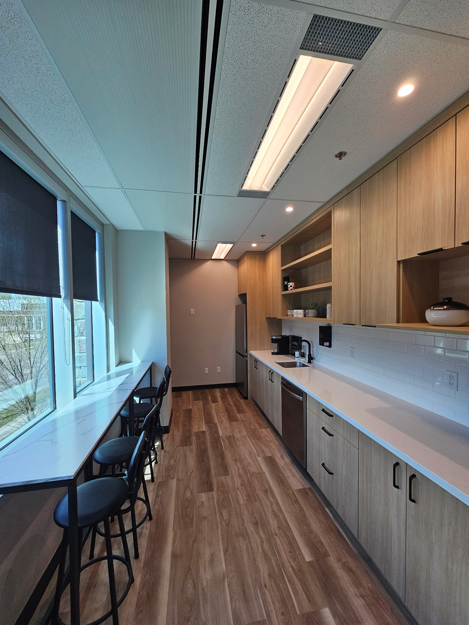  Staff kitchen with warm wood cabinetry, white quartz counters, subway tile, and window-side bar seating with natural light.