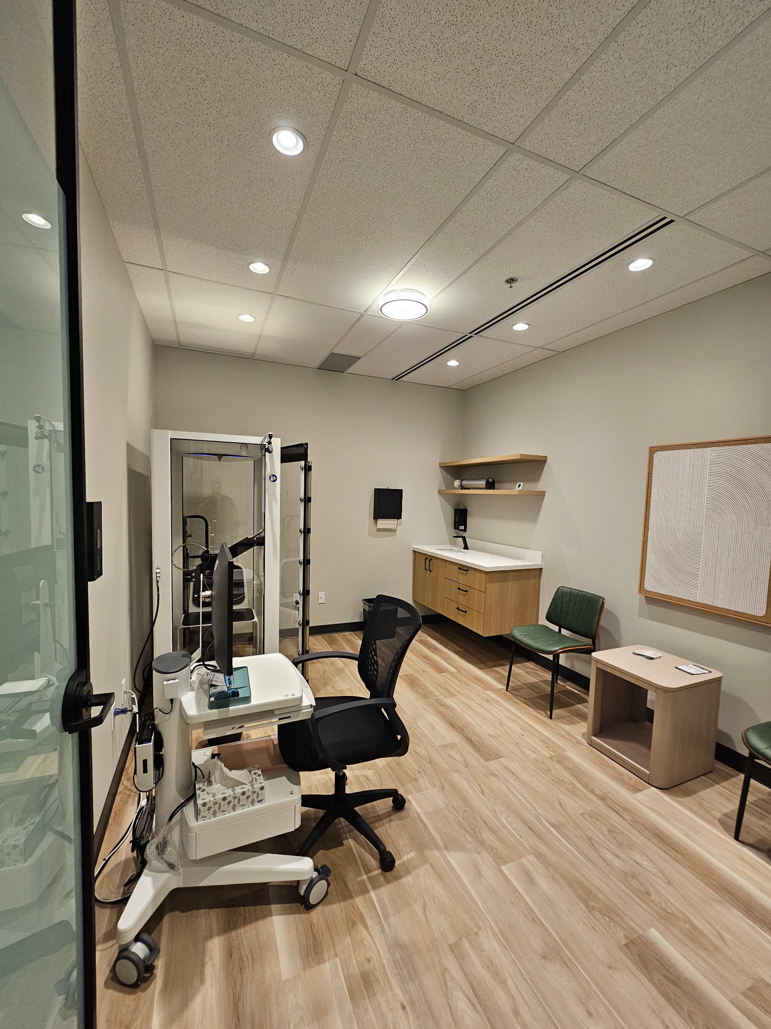 Consulting room with pulmonary testing chamber, floating wood vanity, green patient chair, and abstract framed artwork.