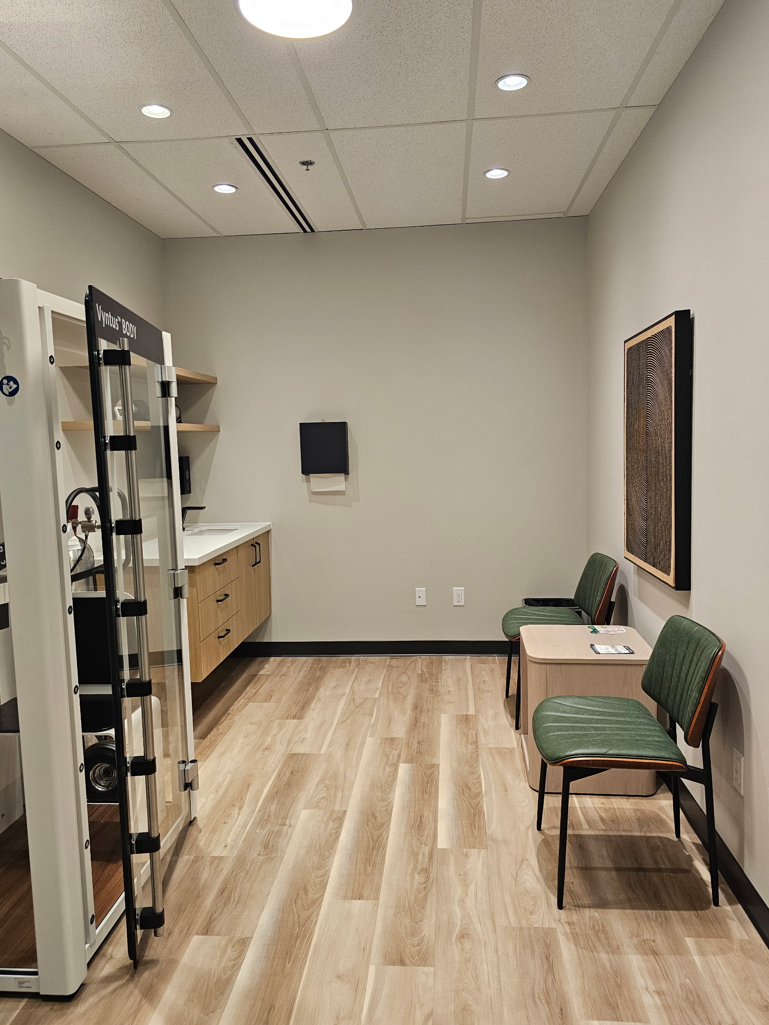 Consulting room with Vyntus Body testing chamber, floating wood vanity, green patient seating, and textured framed artwork.