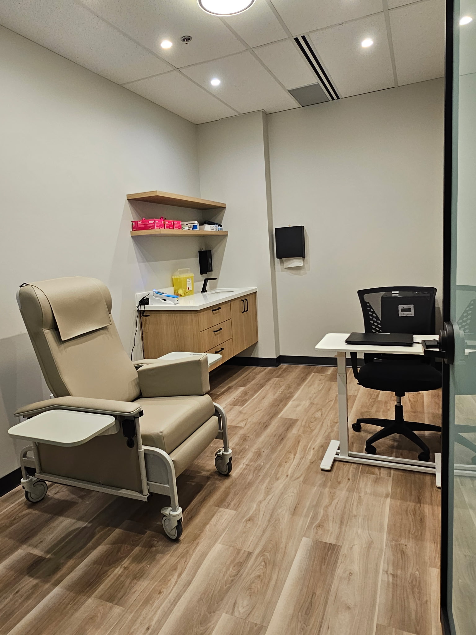 Consulting room with clinical recliner, floating corner vanity, height-adjustable clinician desk, and glazed partition wall.