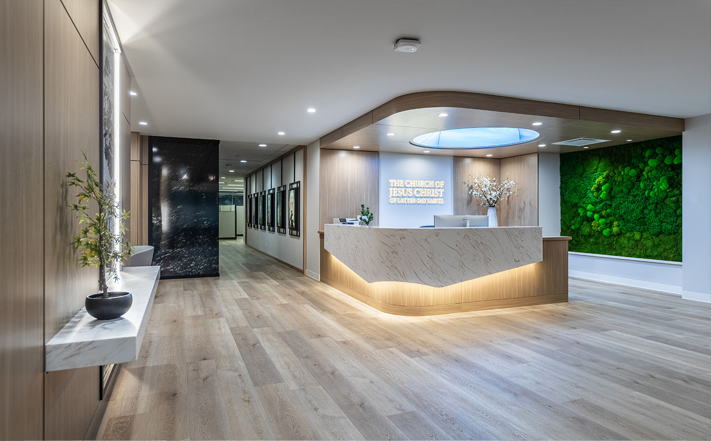 Wide-angle reception with curved marble desk, moss wall, skylight, and gallery hallway leading to offices.