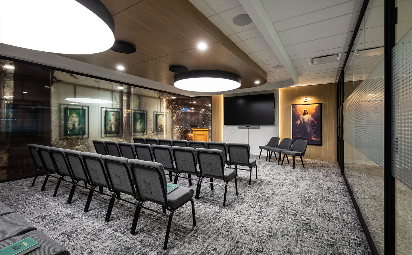 Conference room with rows of gray chairs, large pendant lights, wood ceiling panels, and religious artwork.