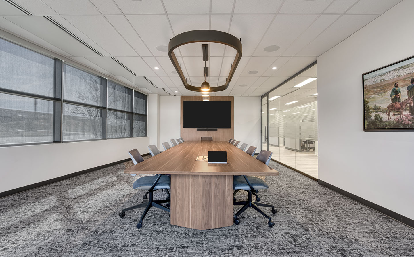 Conference room with boat-shaped wood table, upholstered chairs, geometric pendant light, and wall-mounted TV.