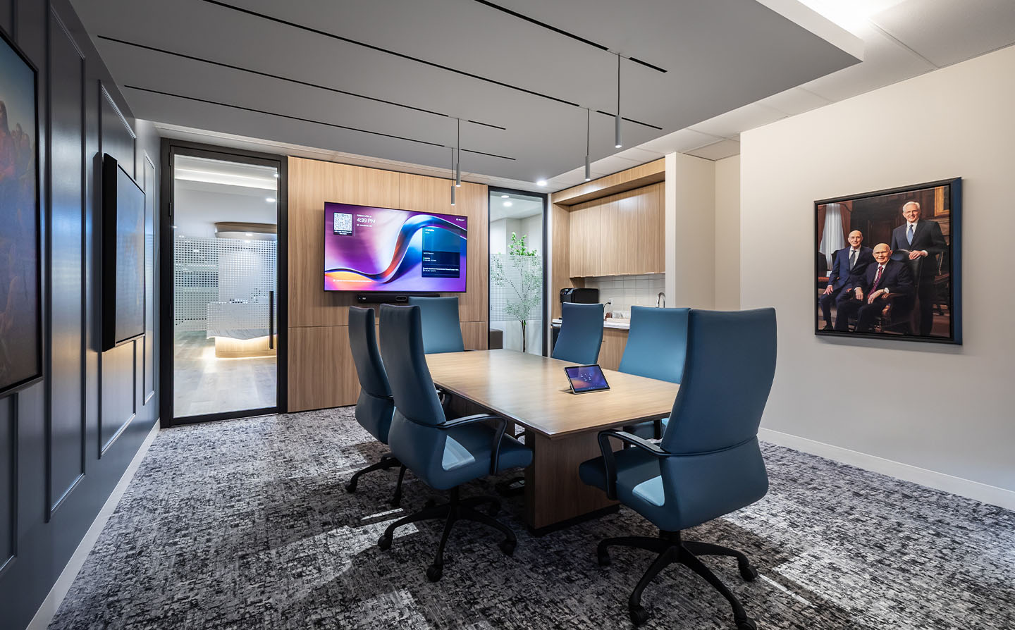 Small meeting room with blue upholstered chairs, wall-mounted TV, wood accents, and portrait photograph on wall.
