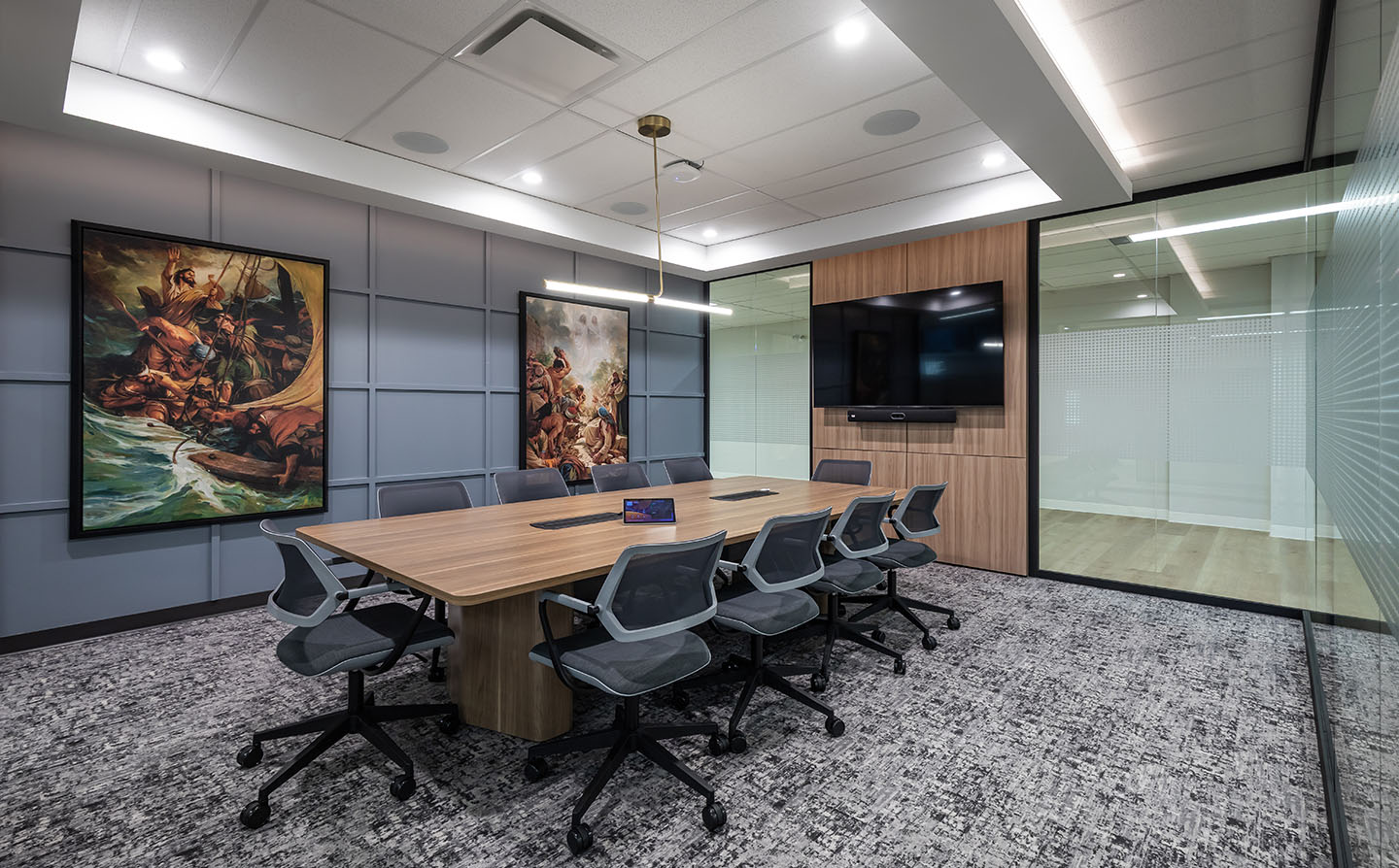 Meeting room with wood conference table, mesh chairs, two large religious paintings, and glass partition wall.