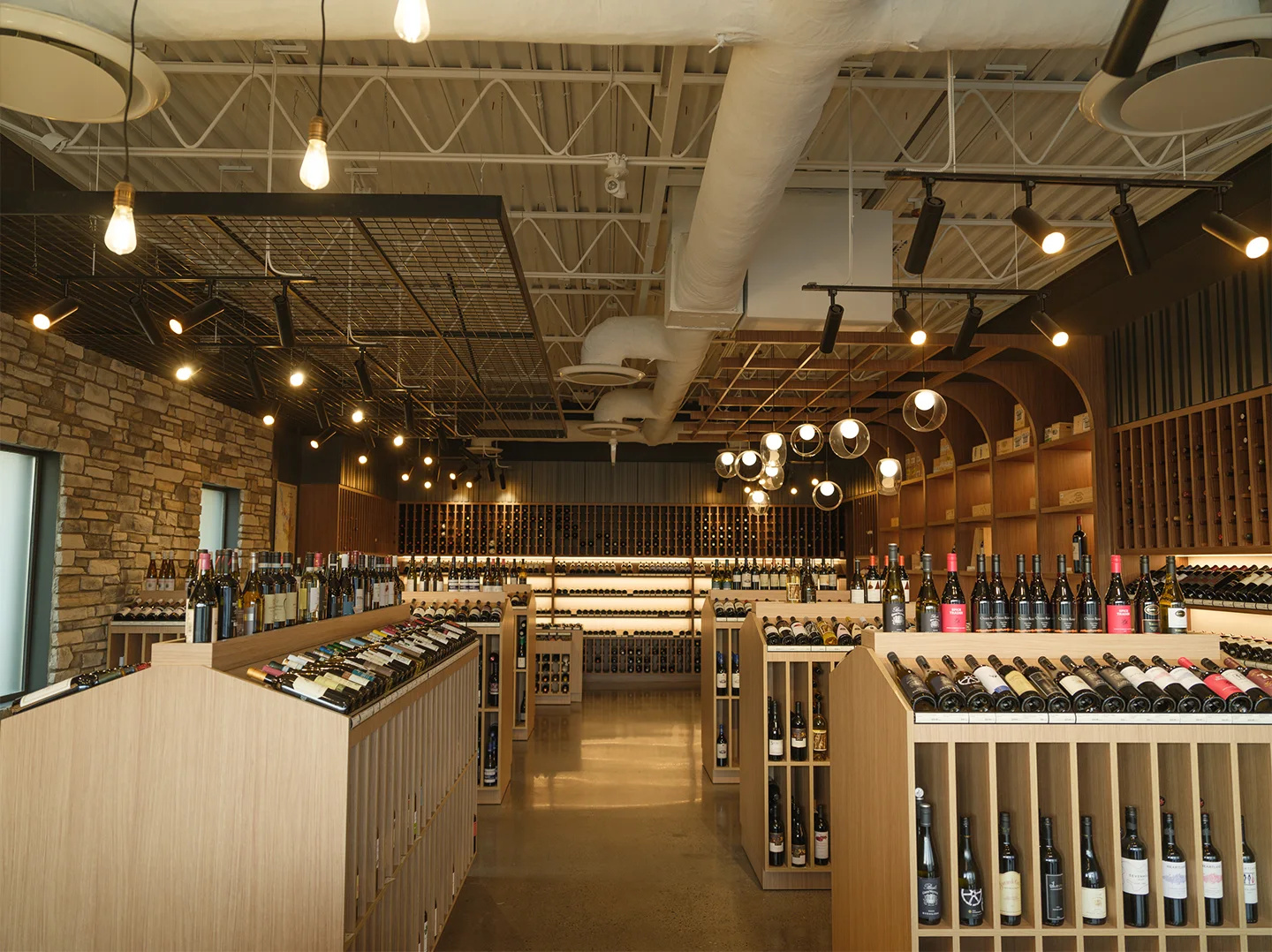 Wide view of the Richmond Hill Wines retail floor, with pale oak display units, bottles angled on top, exposed ceiling ductwork, track lighting, and a stone feature wall.