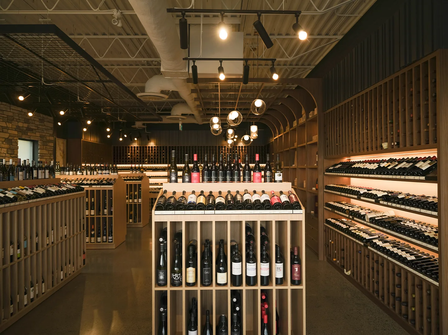 Central aisle of the wine shop with an oak island display in the foreground and a warmly backlit wall of angled bottle shelving on the right.