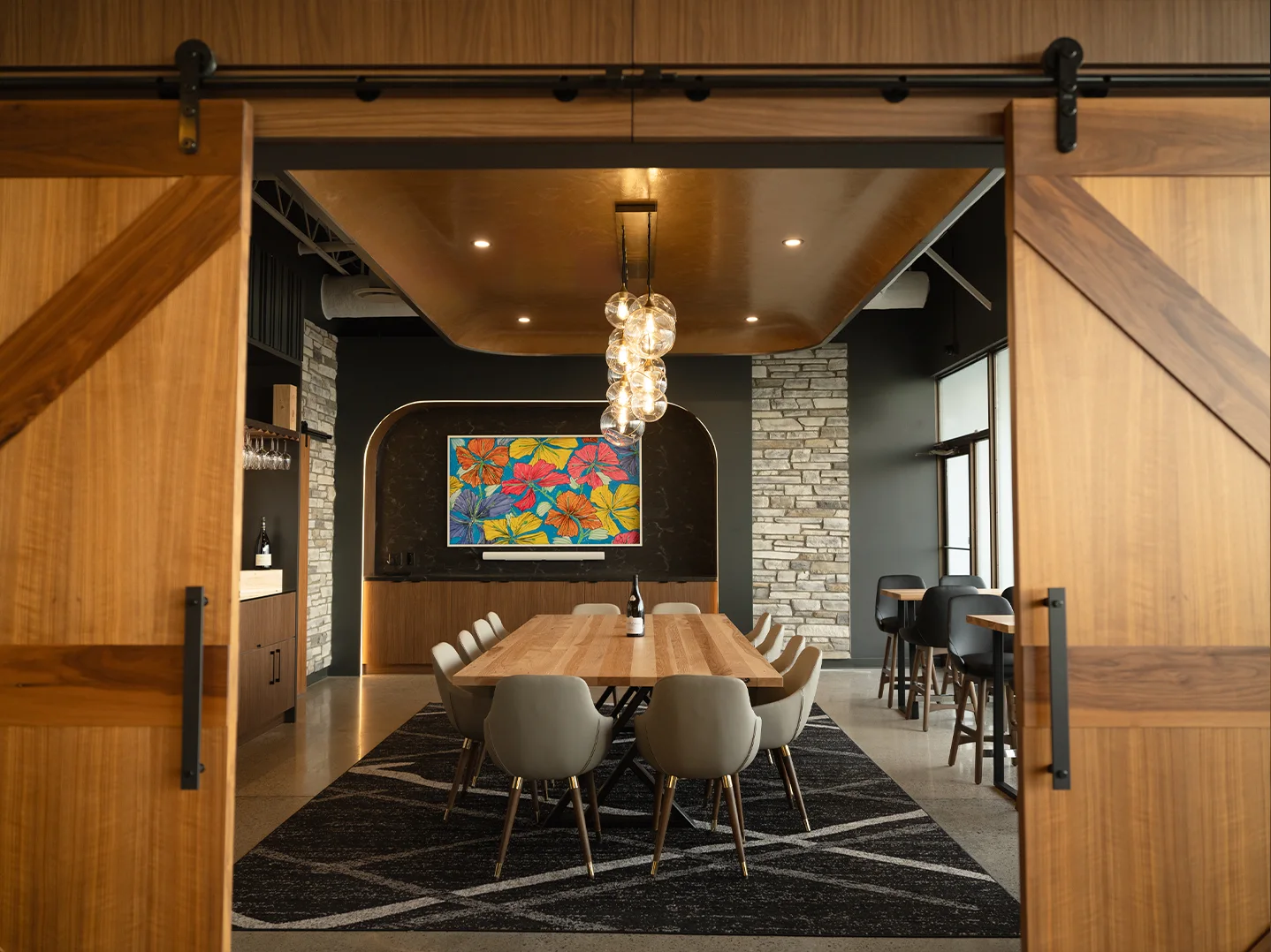 View through open walnut barn doors into the private tasting room, with a long oak table, upholstered chairs, a colourful floral painting, and a cluster of glass pendant lights.