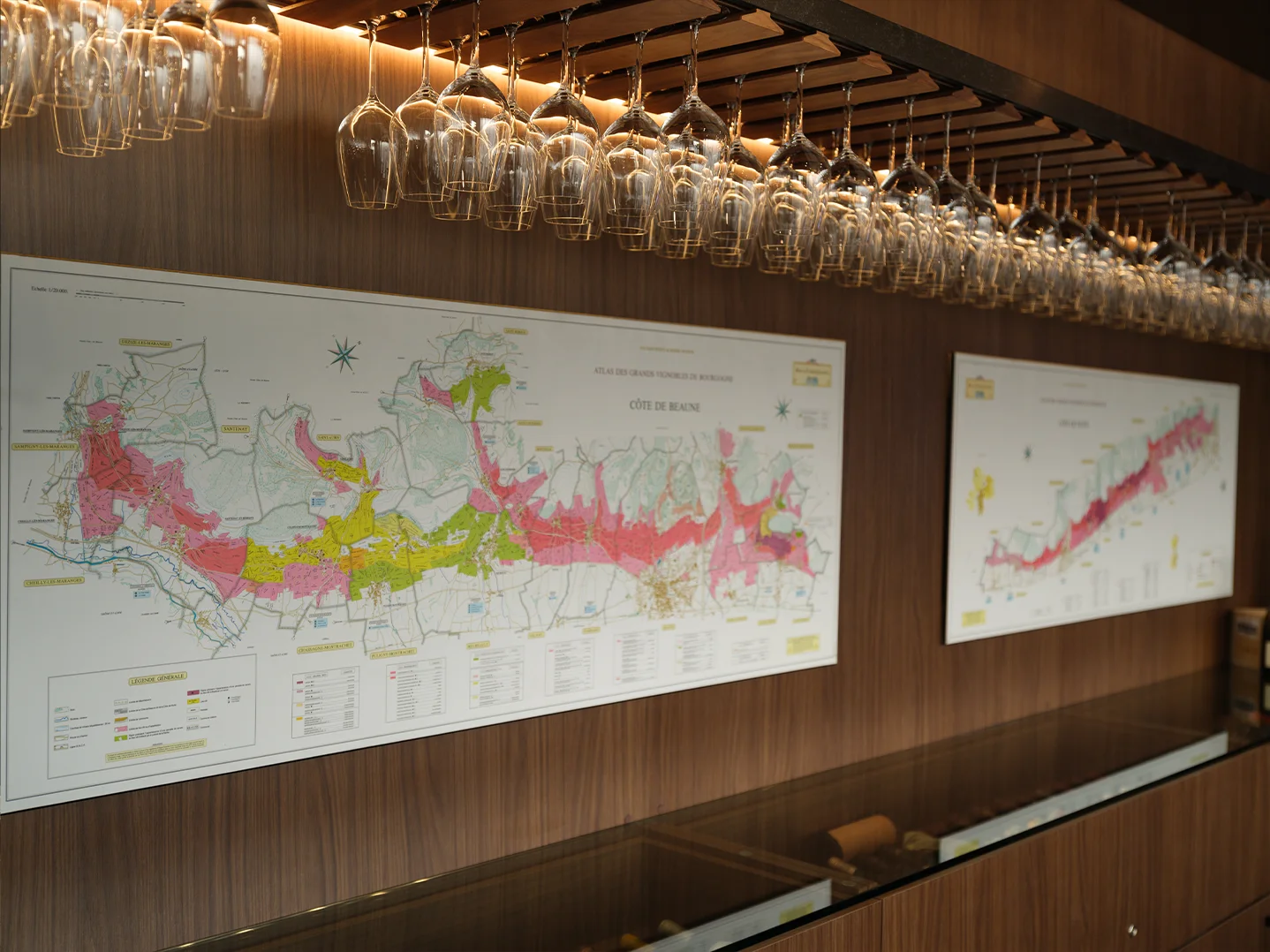 Close-up of the Côte de Beaune map beside a row of hanging stemware lit by warm LED strips, with a glass-topped walnut credenza below.