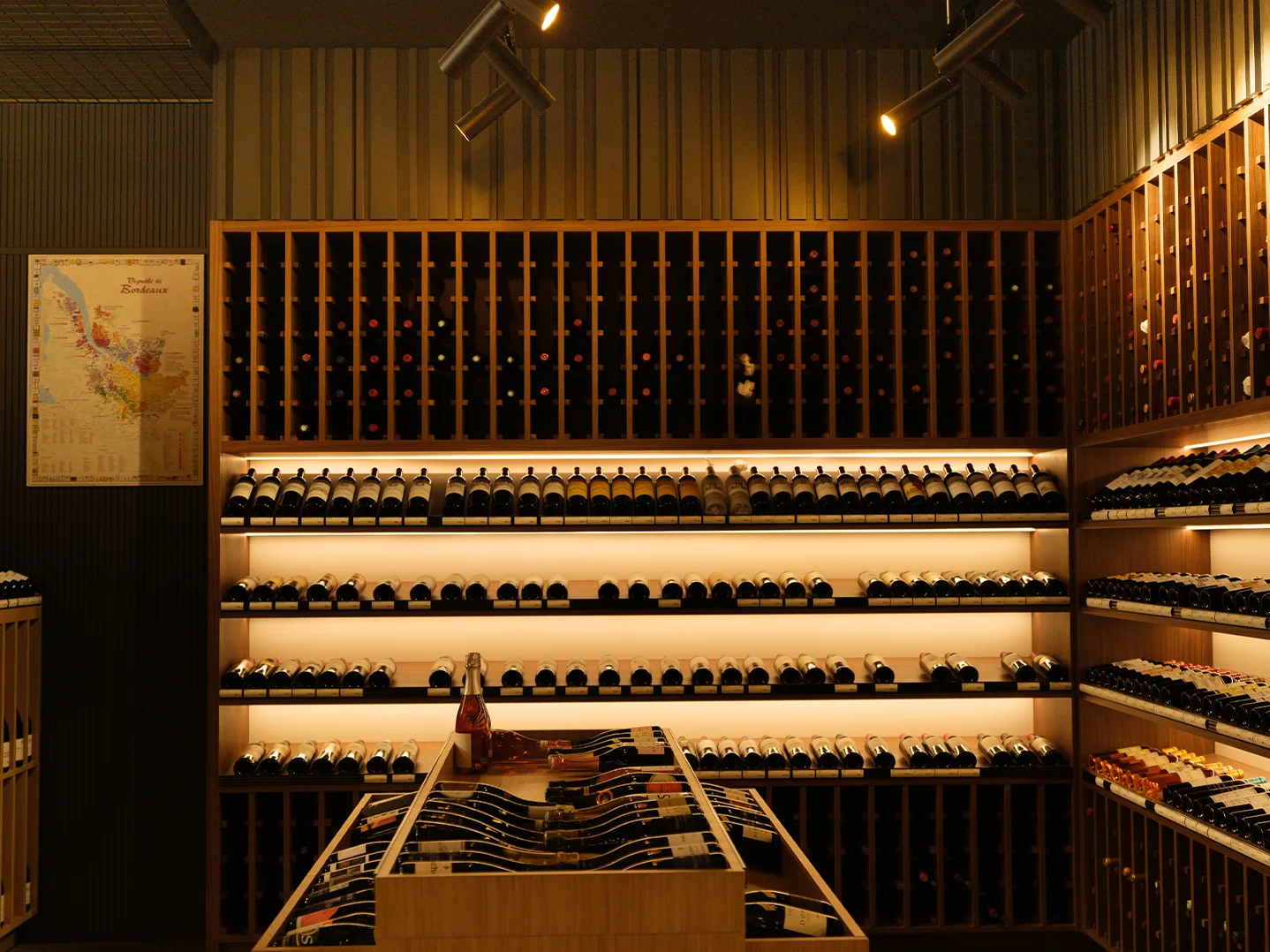 Bordeaux wall with rack storage above and backlit display shelves below, a framed Bordeaux map on the left, and a rosé sparkling bottle on a display table in the foreground.