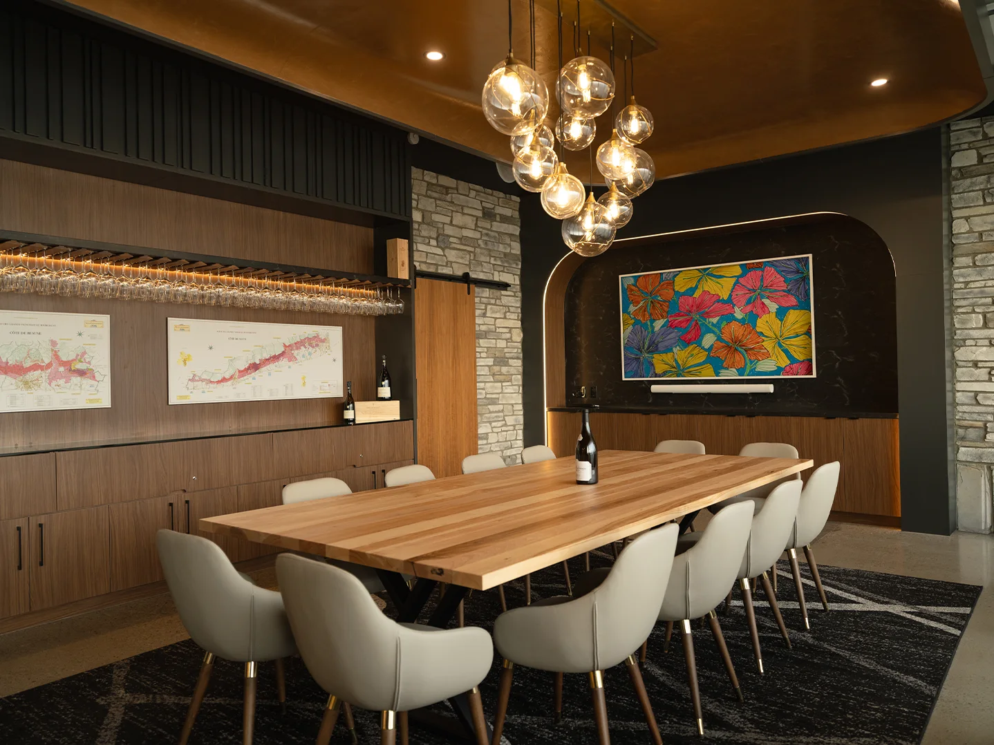 Angled view of the tasting room showing glass globe pendant lights above the oak table, with Burgundy wine region maps and hanging stemware along the left wall.