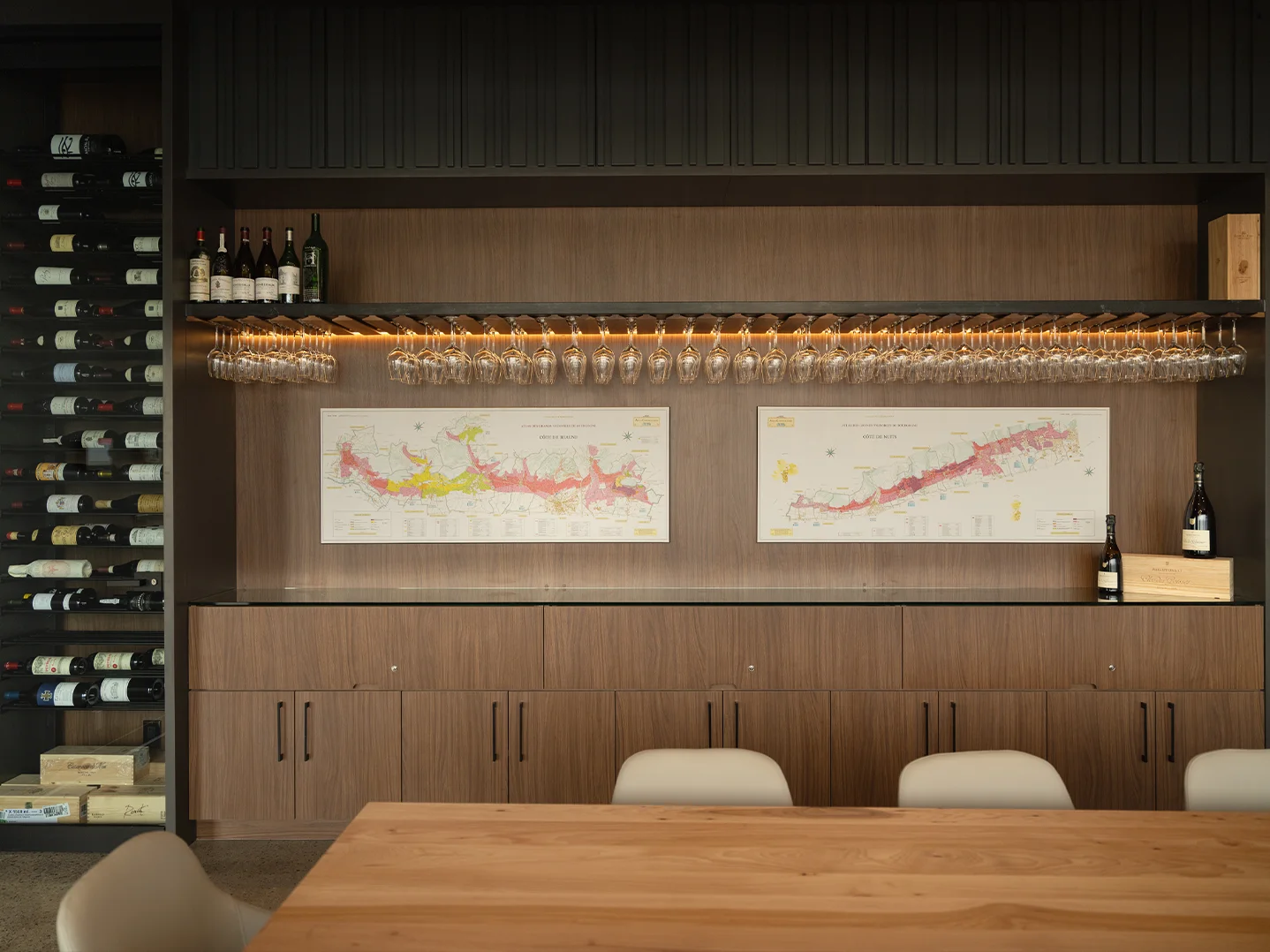Two framed Burgundy region maps, Côte de Beaune and Côte de Nuits, mounted on a walnut wall below a LED-lit rack of hanging wine glasses.