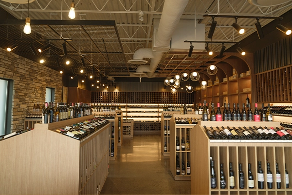 Wide view of the Richmond Hill Wines retail floor, with pale oak display units, bottles angled on top, exposed ceiling ductwork, track lighting, and a stone feature wall.