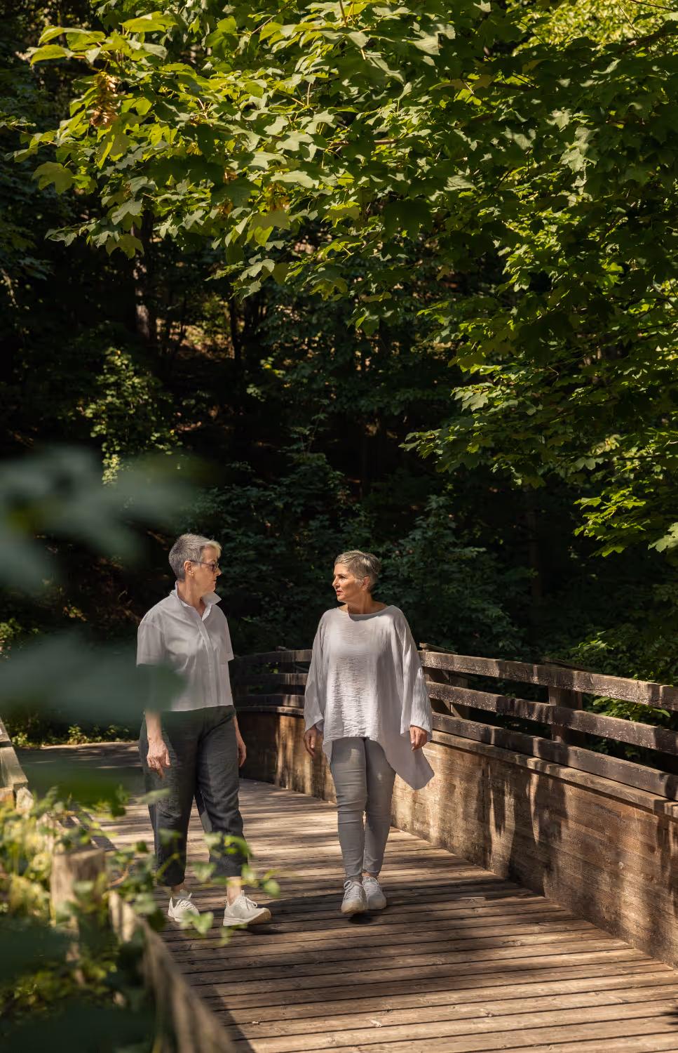 Sylvia und Angelika gehen auf einer Brücke in der Natur und unterhalten sich