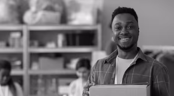 Smiling man holding a cardboard box in a room with shelves and people working in the background.