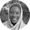 Smiling young woman wearing a light-colored headscarf outdoors with blurred foliage background.