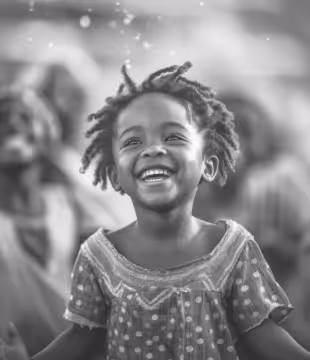 Smiling young girl with curly hair wearing a patterned dress looking upward with blurred people in background.