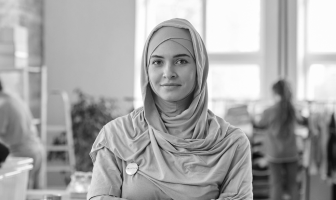 Portrait of a woman wearing a hijab standing indoors with a blurred background of people and furniture.