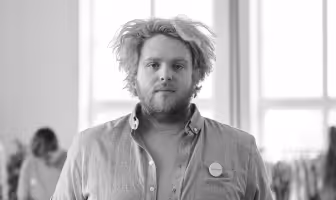 Man with tousled hair and beard wearing an open button-up shirt and button badge, standing indoors near large windows.
