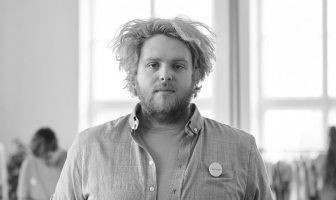 Man with tousled hair and beard wearing an open button-up shirt and button badge, standing indoors near large windows.