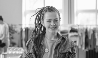 Smiling young woman with dreadlocks wearing a casual shirt in a bright room with clothing racks in the background.