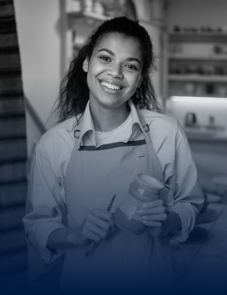 Smiling woman wearing an apron holding a paintbrush and a jar of paint in a workshop.