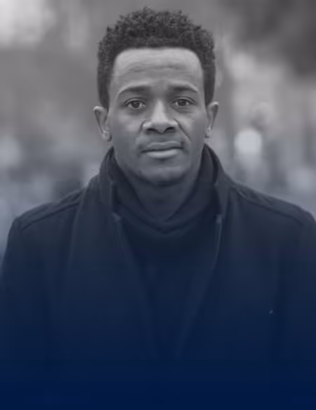 Portrait of a young man with short curly hair wearing a dark coat and scarf, looking directly at the camera outdoors.